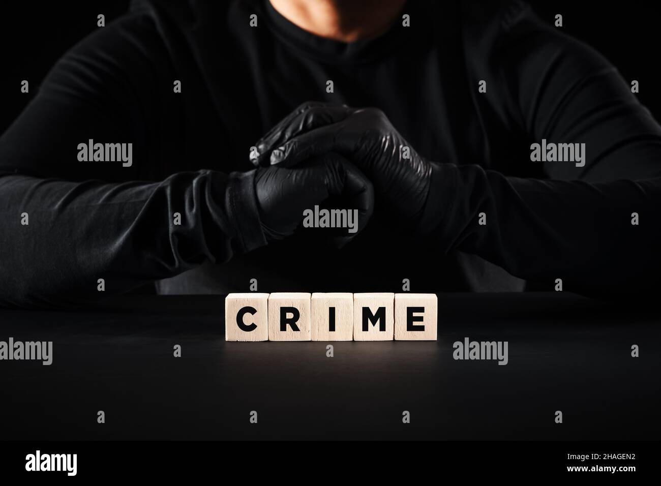 Crime, criminal or criminality concept. Criminal wearing black gloves with clasped hands sits behind the wooden blocks with the word crime. Stock Photo
