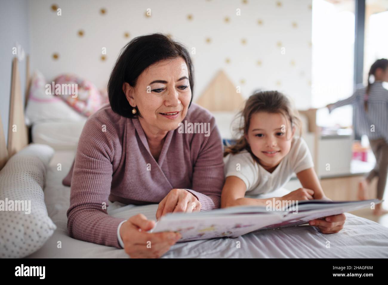 Happy small girl with senior grandmother indoors in bedroom at home, reading on bed. Stock Photo