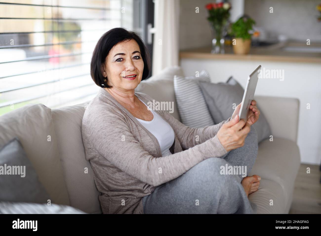 Happy senior woman with tablet sitting on sofa and looking at camera indoors at home, video call concept. Stock Photo
