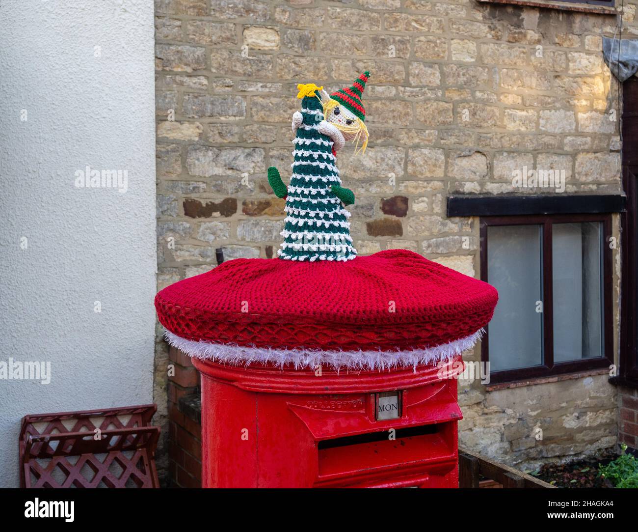 Red Royal Mail post box topped with a knitted Christmas decoration, or yarn bombing, in the village of Hackleton, Northamptonshire, UK Stock Photo