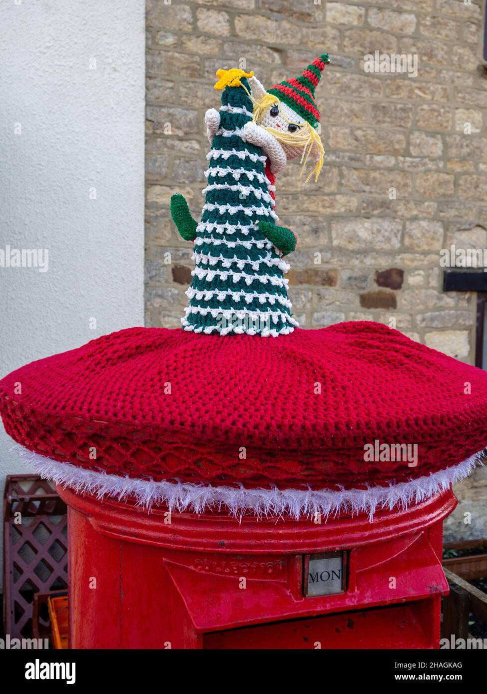 Red Royal Mail post box topped with a knitted Christmas decoration, or yarn bombing, in the village of Hackleton, Northamptonshire, UK Stock Photo
