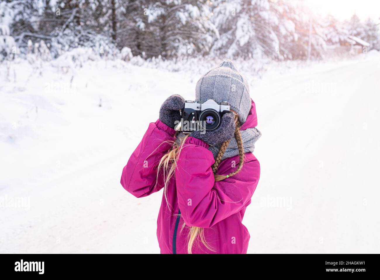Backlit portrait of girl in warm clothes taking photo in winter forest. Child with camera for walk in winter. Day of photographer. Hobby, photography Stock Photo