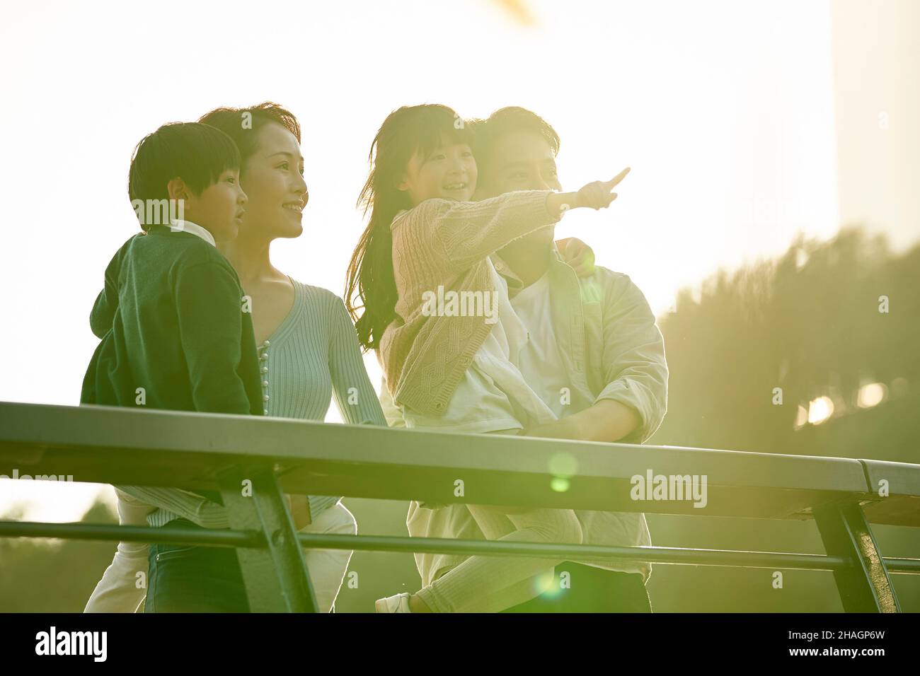 asian family with two children standing on pedestrian bridge looking at view in city park at sunset Stock Photo
