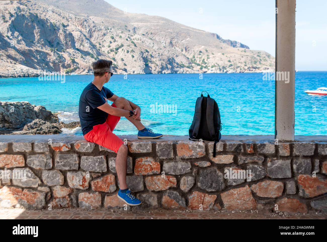 A man in red shorts sits on a stone fence and looks out over the turquoise sea. Stock Photo