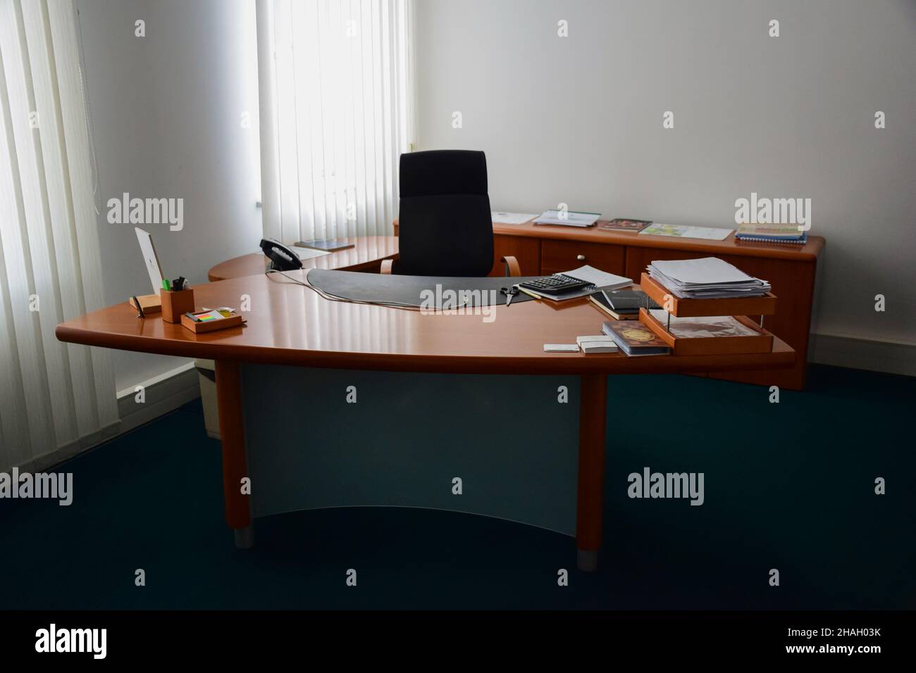 Work office space with an office chair, brown wooden table and various items on it near two windows. Front view Stock Photo