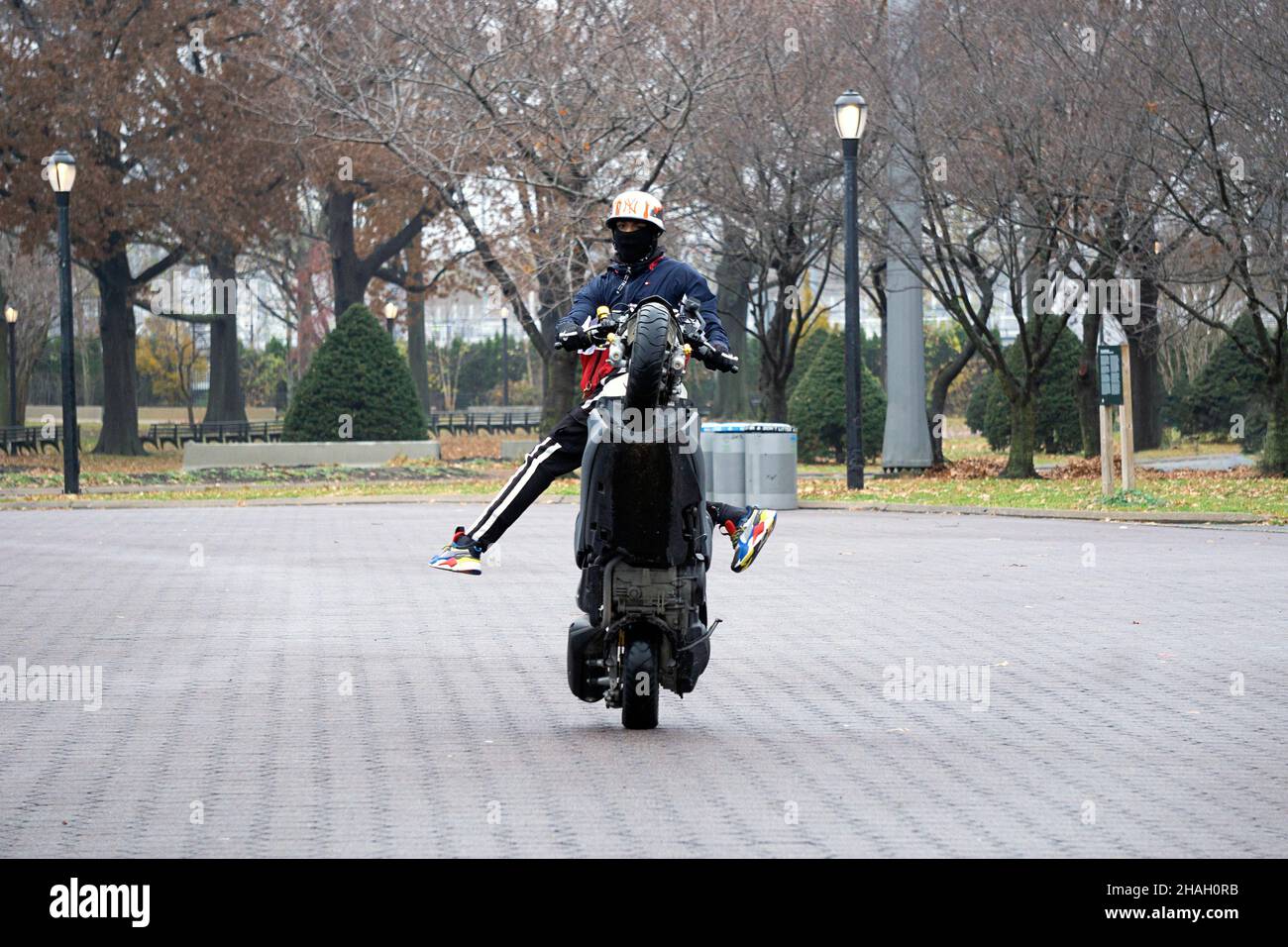 A teenage biker with a helmet & mask does a wheelie on his Yamaha TMax while his legs are off the bike. In a park in Queens, New York City. Stock Photo
