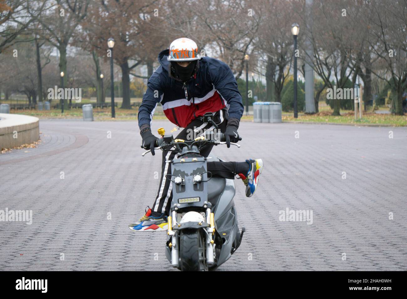 A teenage biker with a helmet & mask rides his Yamaha TMax while his legs are off the bike. In a park in Queens, New York City. Stock Photo