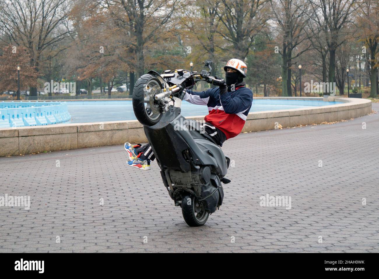 A teenage biker with a helmet & mask does a wheelie on his Yamaha TMax while his legs are off the bike. In a park in Queens, New York City. Stock Photo