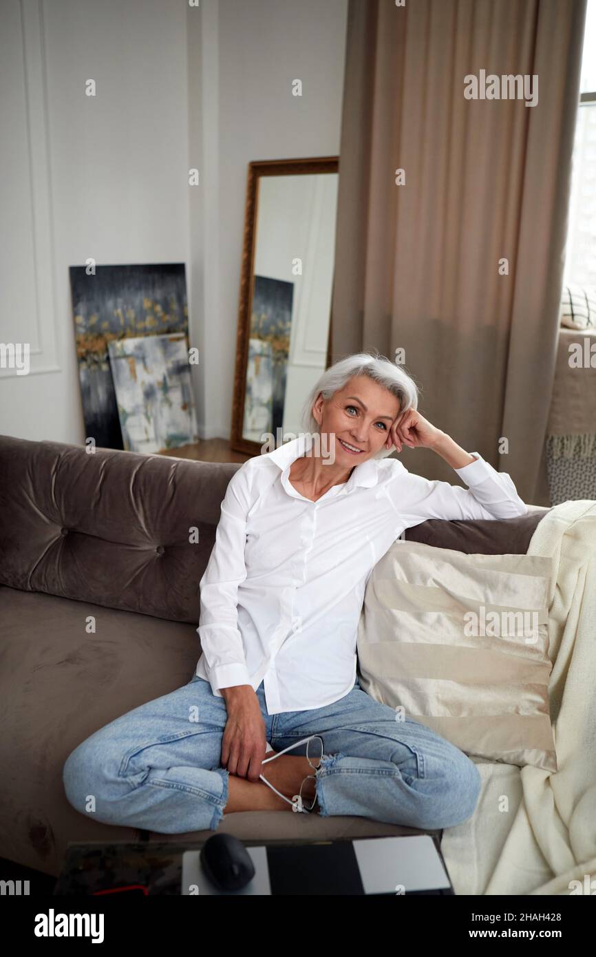 High angle of charming middle aged female with gray hair and in jeans and white shirt sitting with crossed legs on couch at home and smiling at camera Stock Photo
