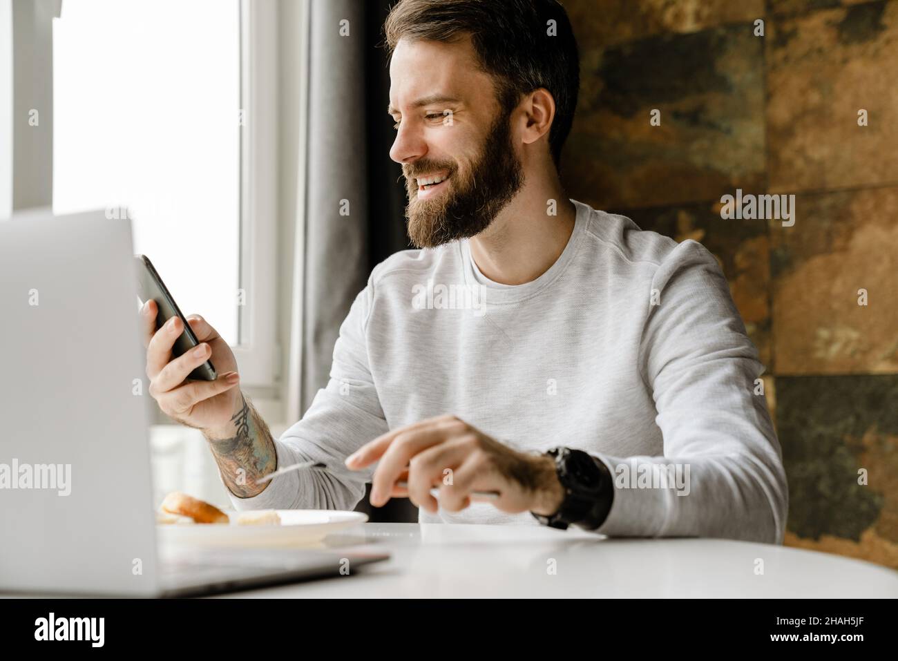 Bearded man smiling and using cellphone while working with laptop indoors Stock Photo