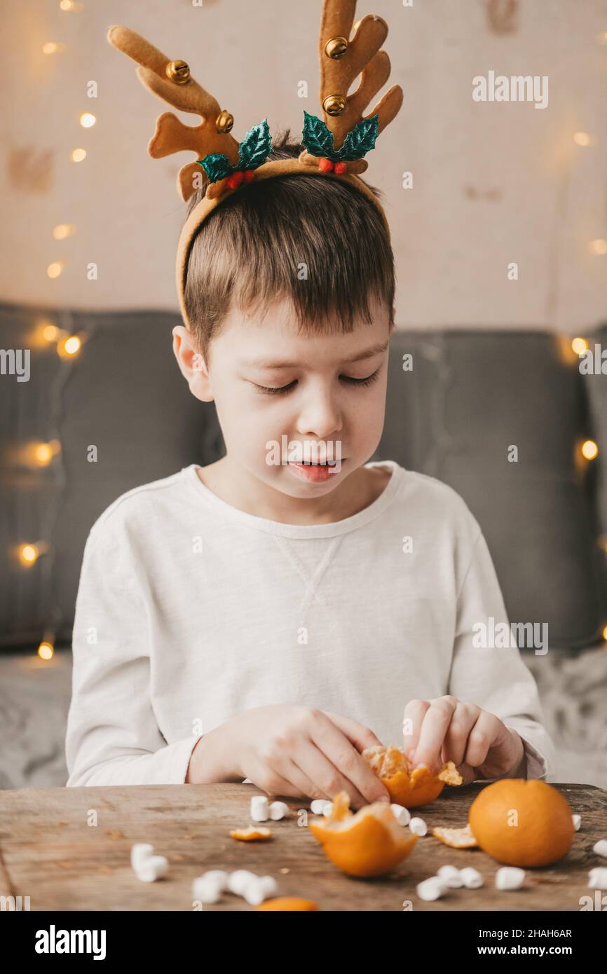 boy in Christmas antlers, sits and peels a tangerine on the background of a yellow garland. Boy in a white t-shirt at home, peels a tangerine Stock Photo