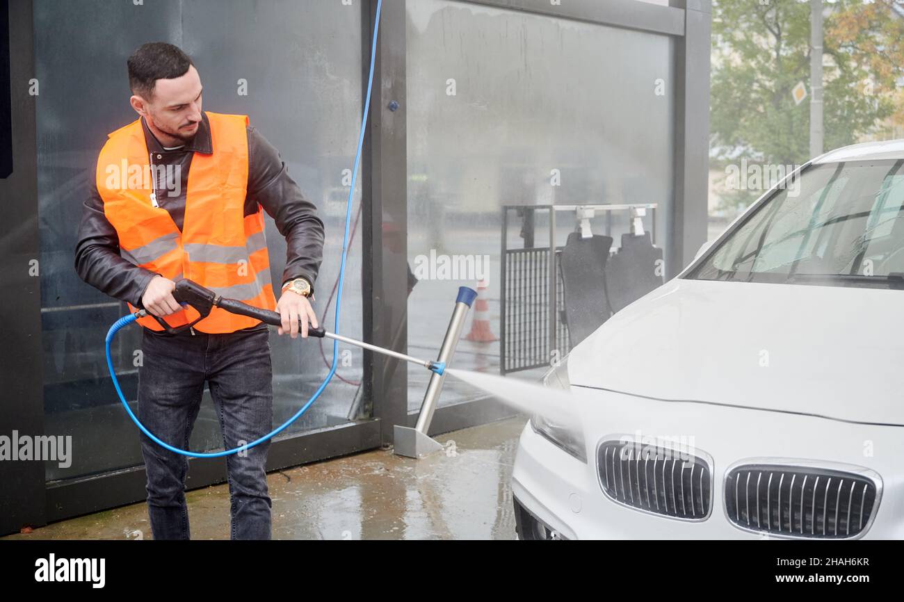 Young man washing car on carwash station, wearing orange vest. Handsome worker cleaning white automobile, using high pressure water. Stock Photo