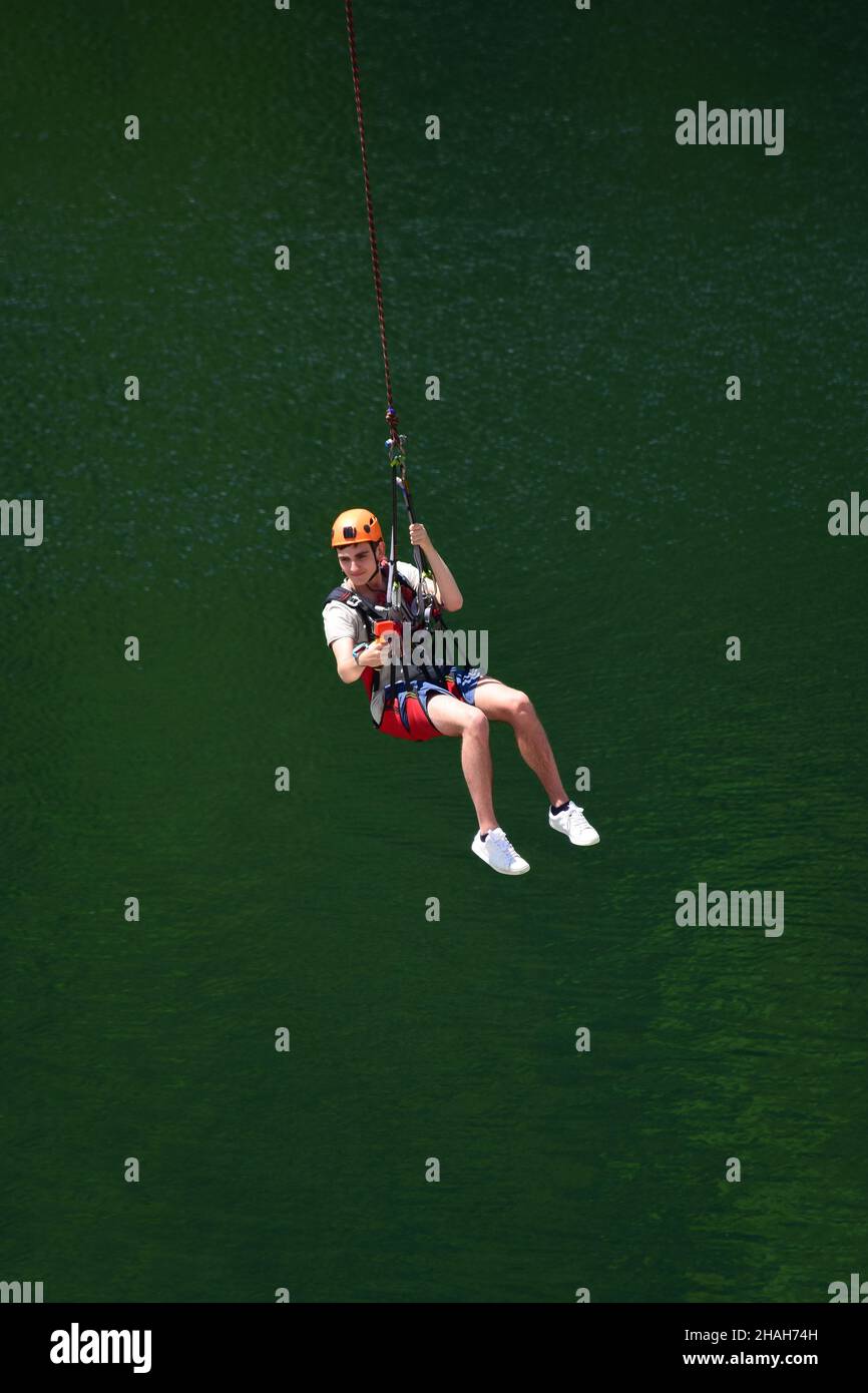 A young man in a helmet jumped from bungee jumping and now hangs on a rope, swinging and filming himself on a sports video camera on a blurred backgro Stock Photo