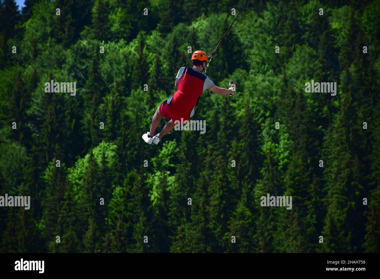 A young man in a helmet jumped from bungee jumping and now hangs on a rope, swinging and filming himself on a sports video camera against a blurred ba Stock Photo