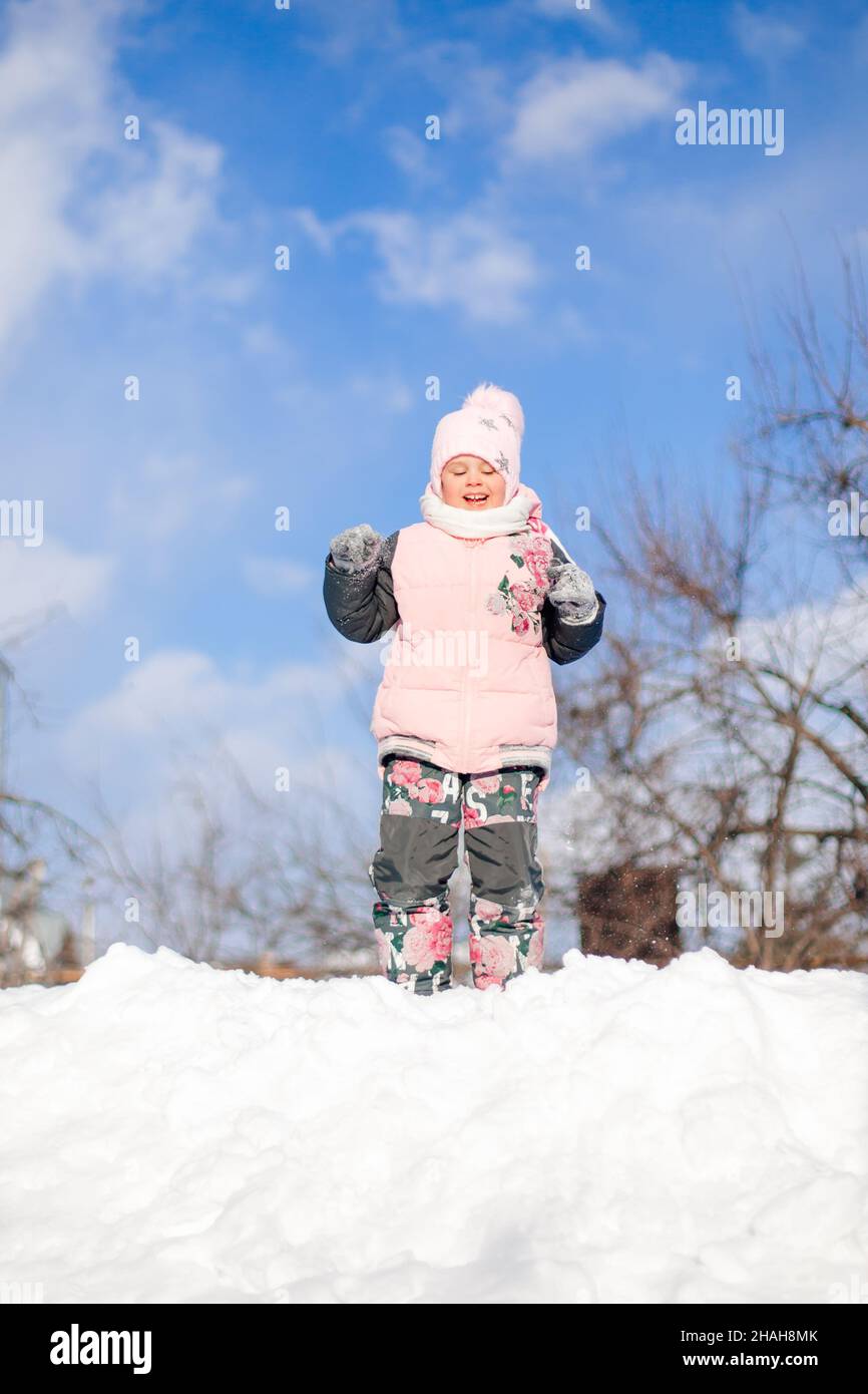Little girl is playing in nature. Child in pink winter jacket and hat with pompom has fun on sunny winter day, plays and rides down snow slide without Stock Photo