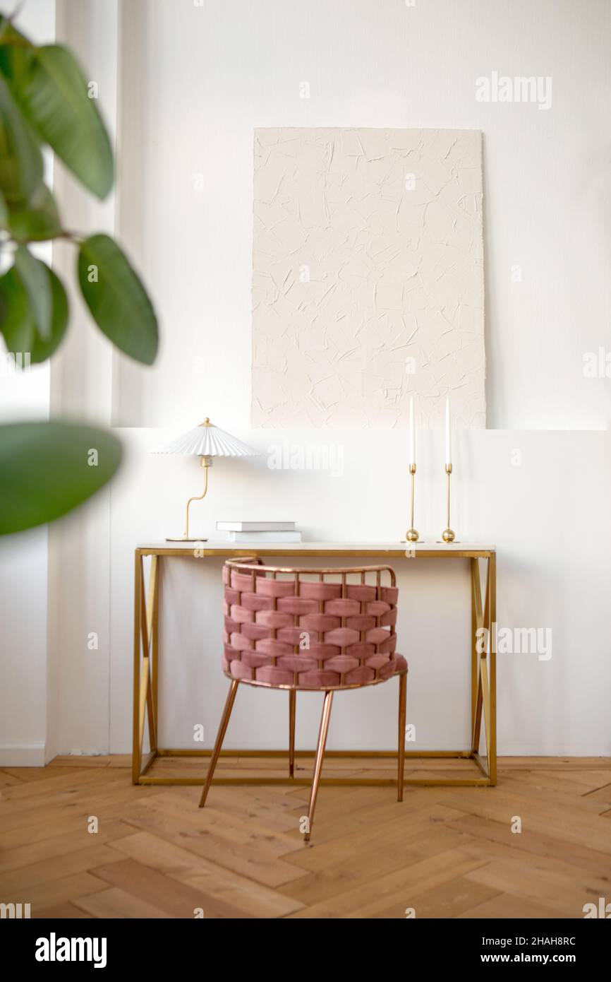 Interior design of light room furnished with table with wicker chair and lamp and books placed near white wall with ledge with white painting on it Stock Photo