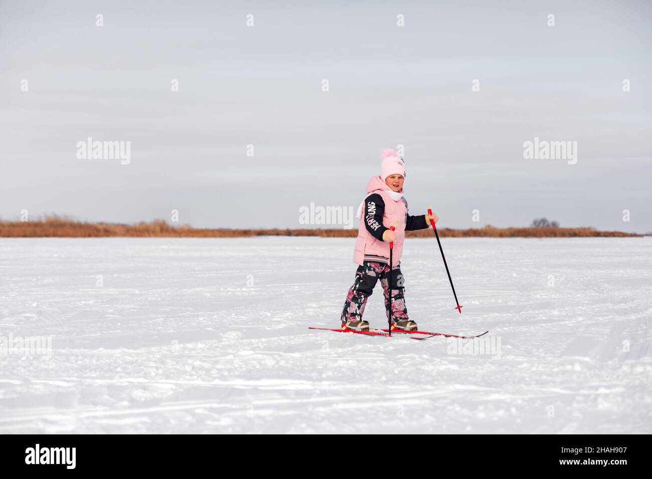 Child learns to ski. Little girl in pink warm suit skiing in snow on frosty winter day, side view, snow background Stock Photo