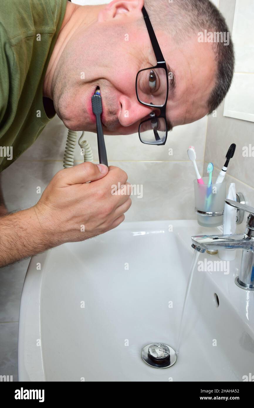 A white man with glasses is brushing his teeth in a bathtub above the sink. The tap is open and water is flowing. Stock Photo