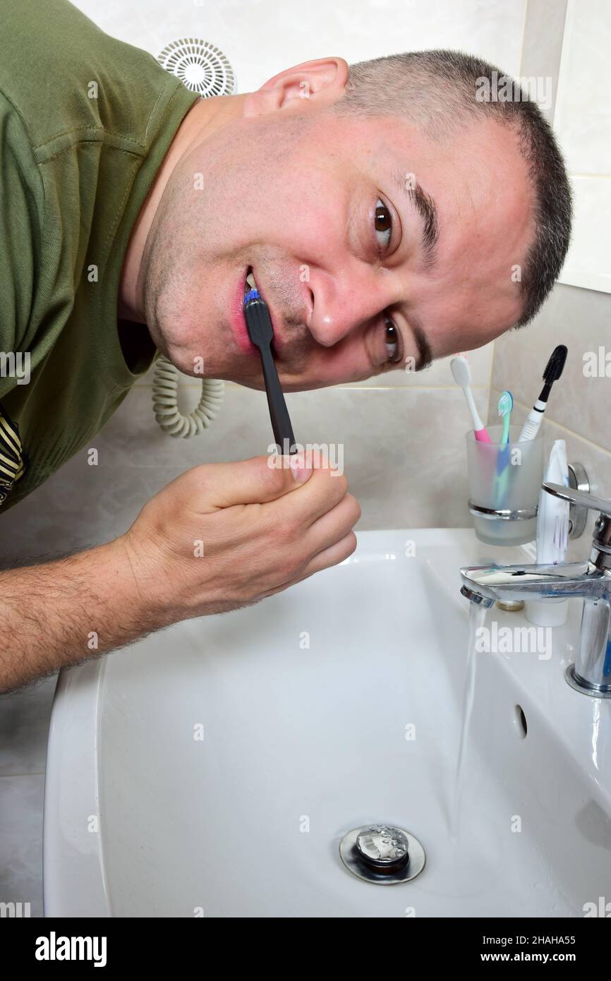 A white man is brushing his teeth in the bathtub above the sink. He looks into the camera. The tap is open and water is flowing. Stock Photo