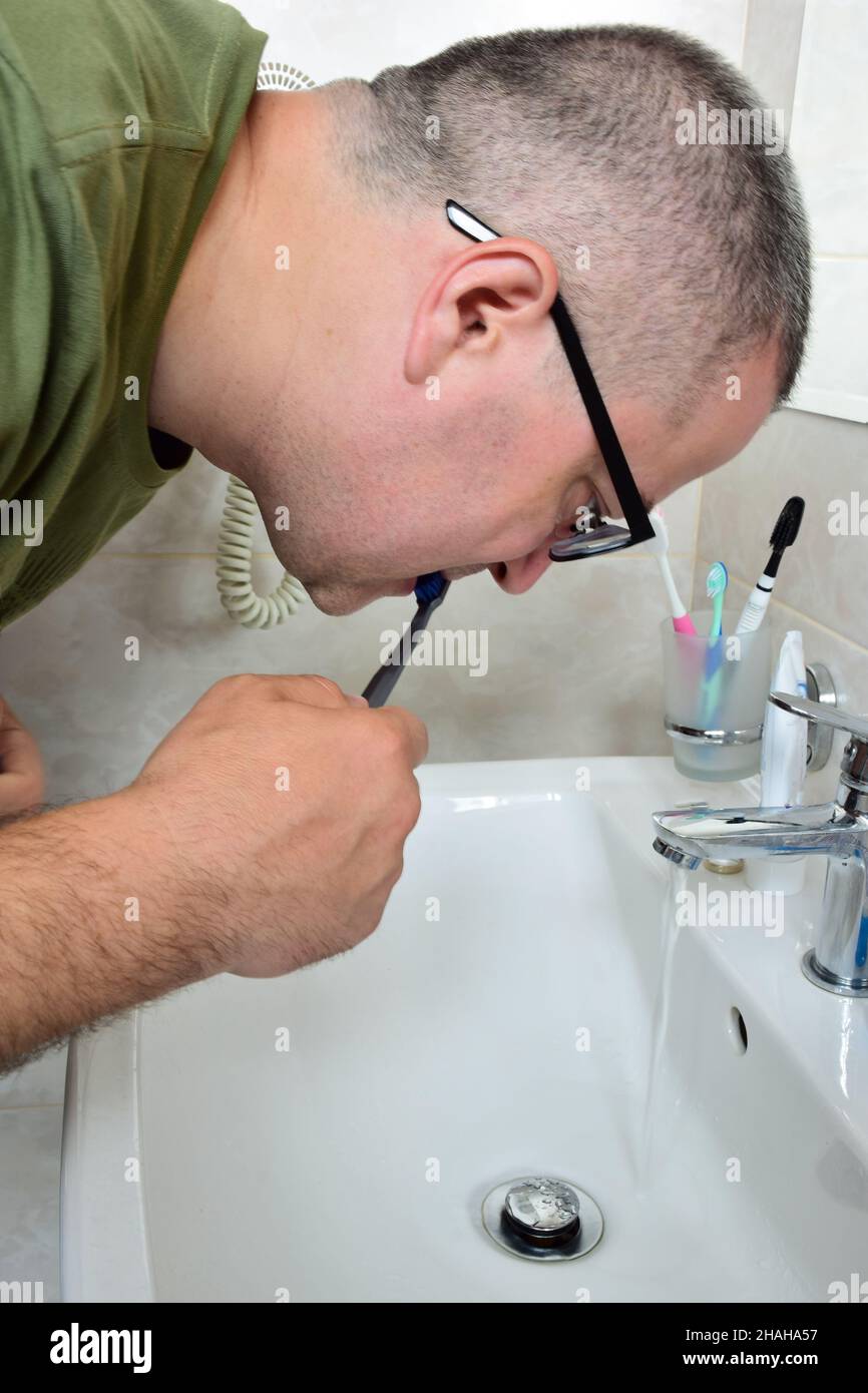 A white man with glasses is brushing his teeth in a bathtub above the sink. He doesn't look at the camera. The tap is open and water is flowing. Stock Photo