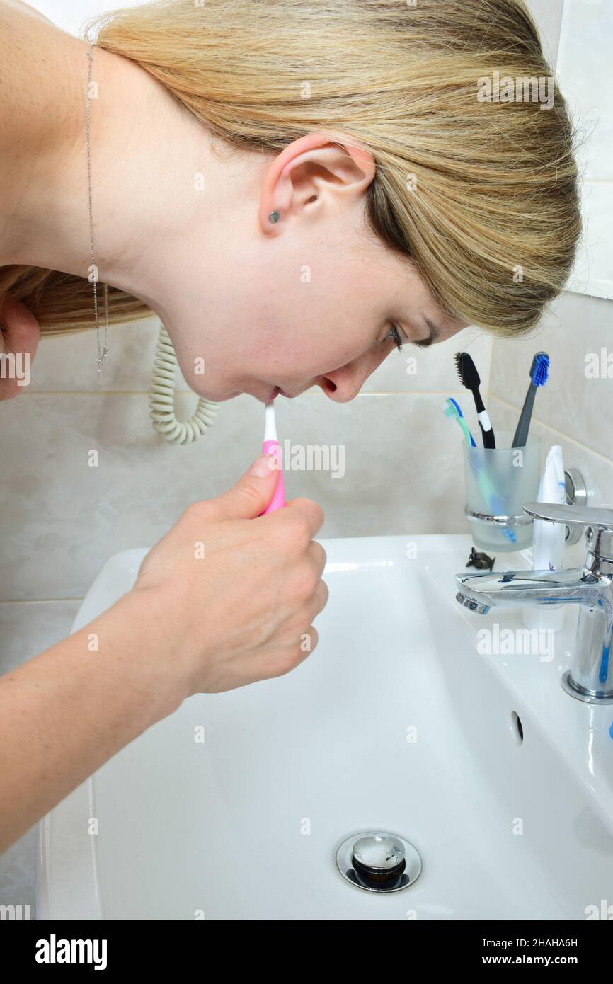 A white woman with long hair is brushing her teeth in a bath above the sink. She doesn't look at the camera. The tap is closed and no water flows. Stock Photo