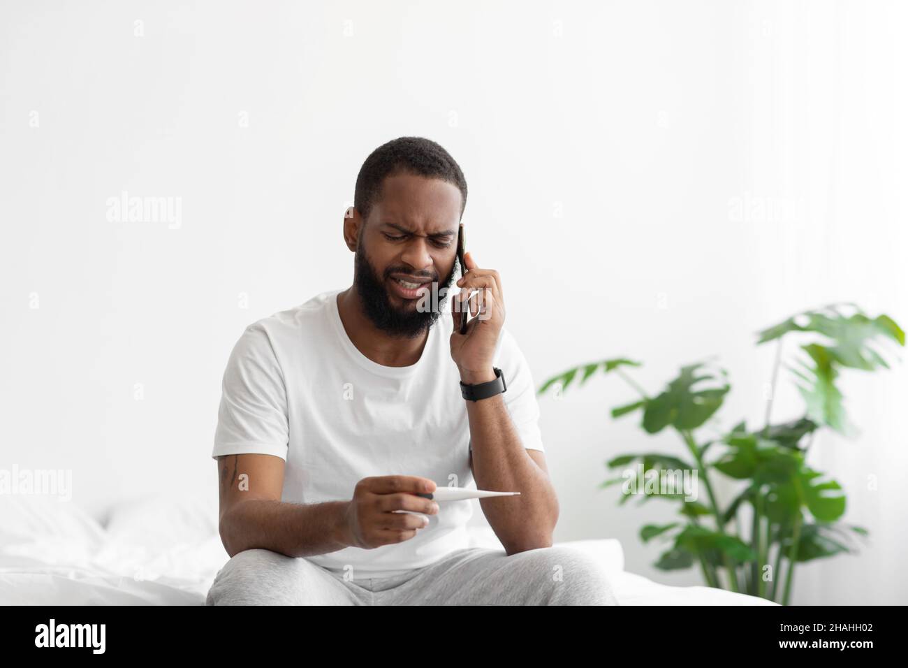 Despaired young black bearded male watch at thermometer with high fever, calling by phone Stock Photo