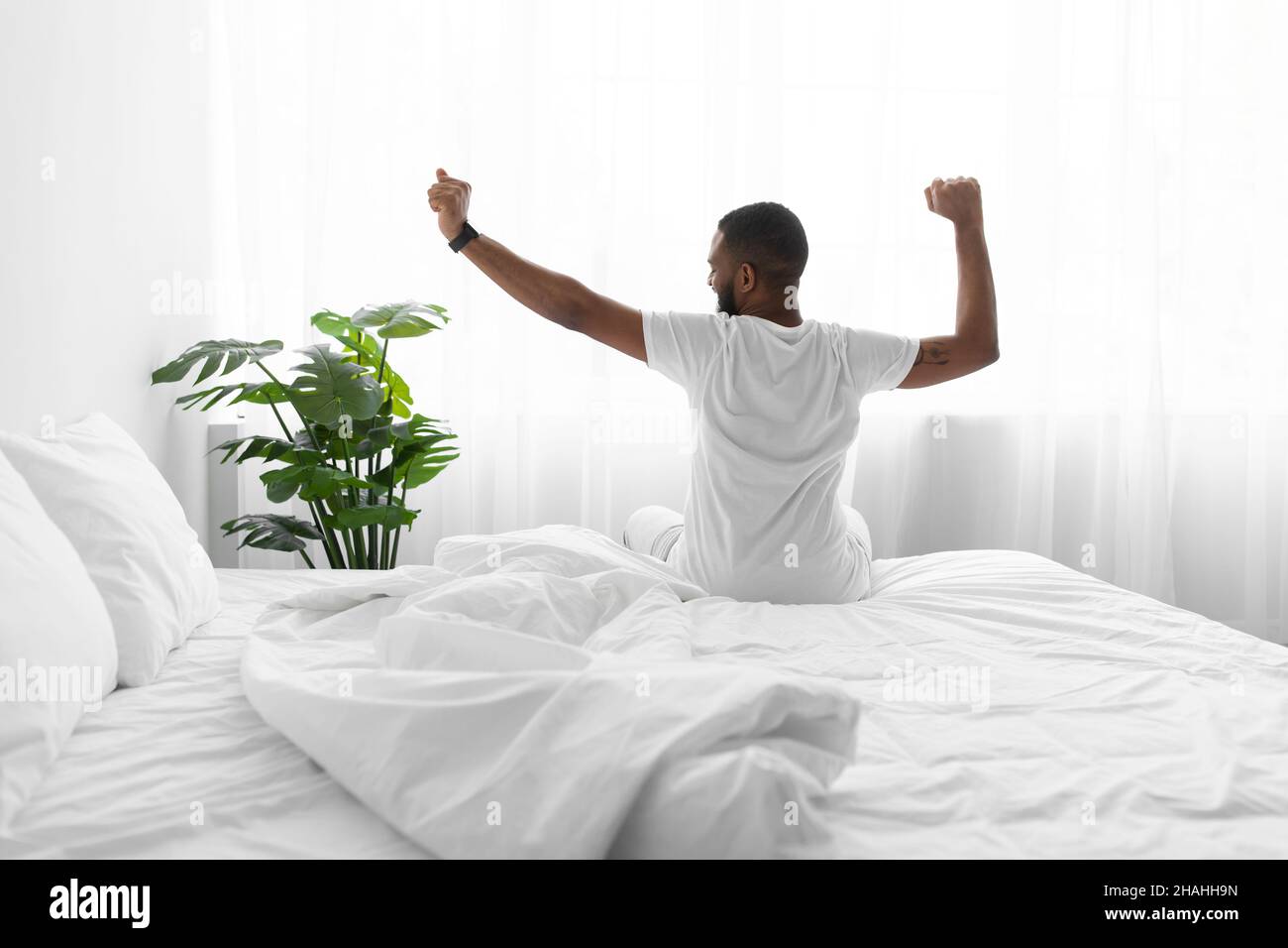 Calm millennial african american guy sitting on bed stretching body and enjoying good morning Stock Photo