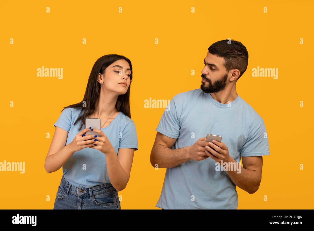 Privacy concept. Arab lady trying to look at her boyfriend's smartphone, guy hiding gadget, yellow studio background Stock Photo