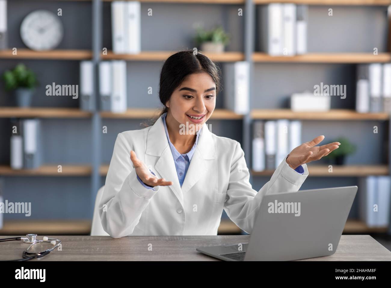 Smiling young indian female doctor in white coat gestures look at ...