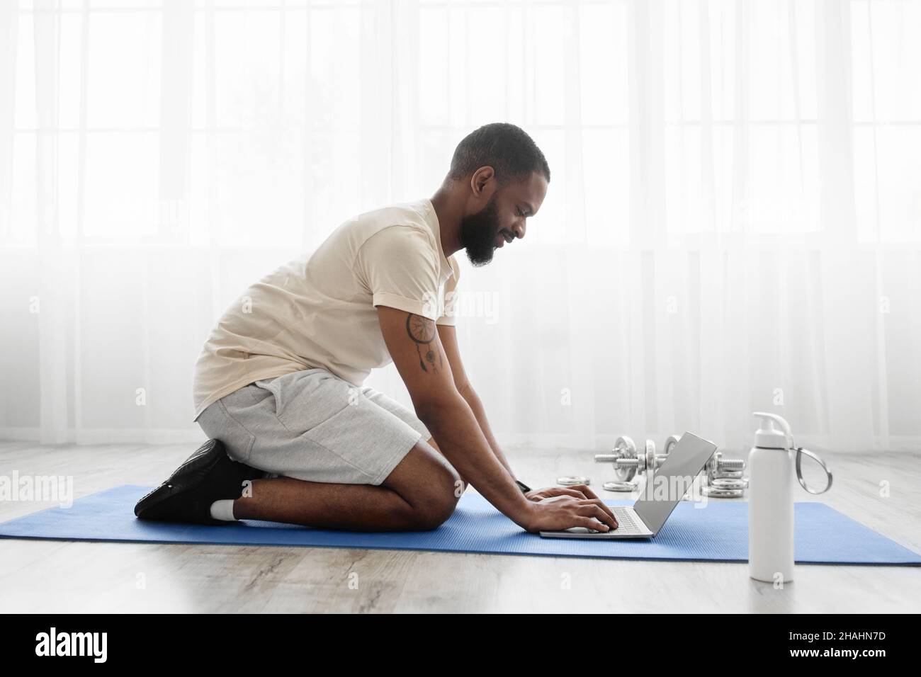 https://c8.alamy.com/comp/2HAHN7D/smiling-young-black-bearded-man-sit-on-mat-on-floor-in-room-with-dumbbells-bottle-of-water-watch-workout-2HAHN7D.jpg