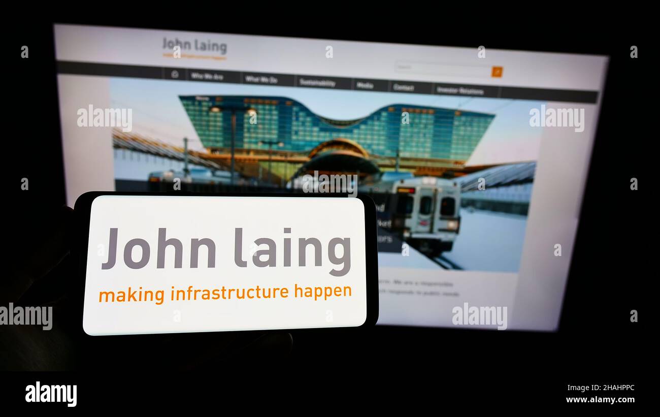 Person holding smartphone with logo of British company John Laing Group Limited on screen in front of website. Focus on phone display. Stock Photo