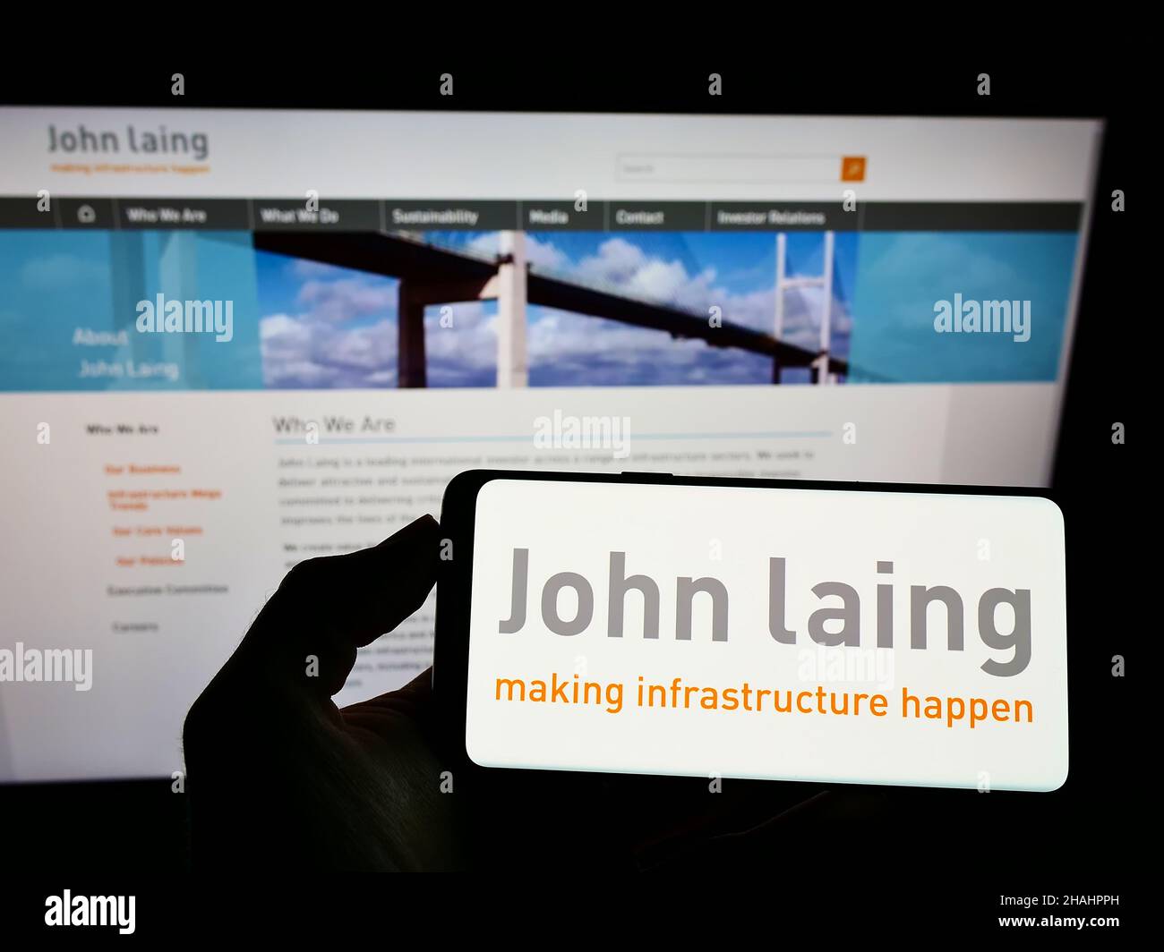Person holding mobile phone with logo of British company John Laing Group Limited on screen in front of business web page. Focus on phone display. Stock Photo