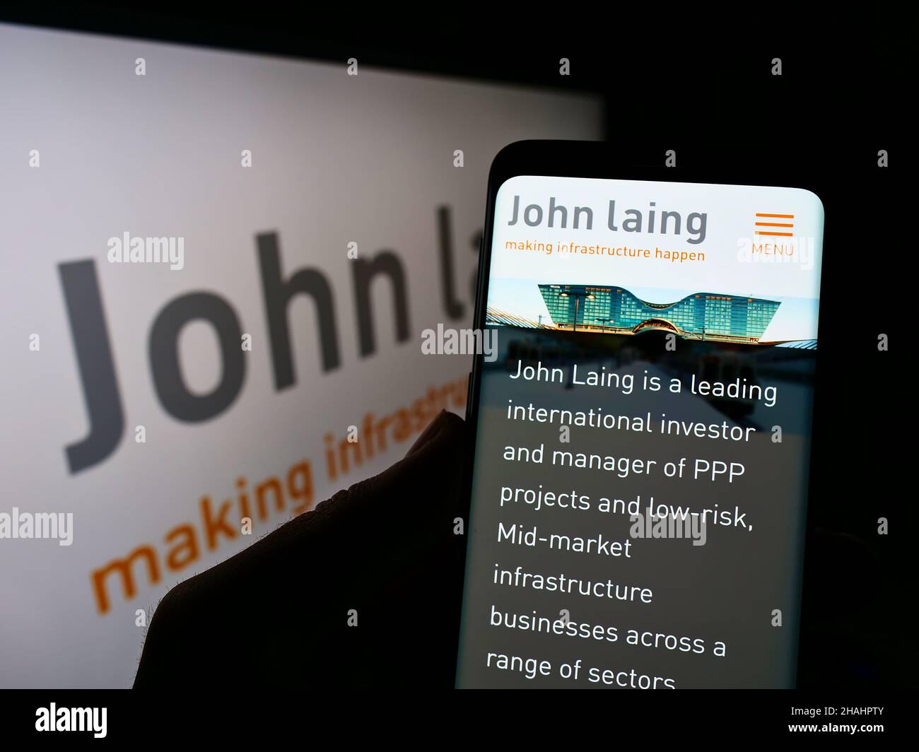 Person holding cellphone with website of British company John Laing Group Limited on screen in front of logo. Focus on center of phone display. Stock Photo
