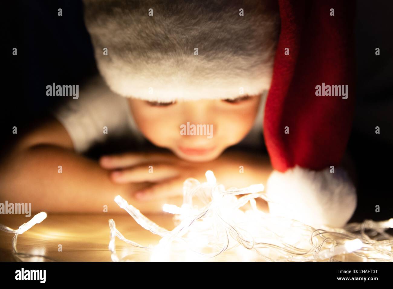 The kid in Santa hat lies on the floor in the dark and plays with Christmas lights. The child imagines magic and a fairy tale. He has a gentle smile Stock Photo