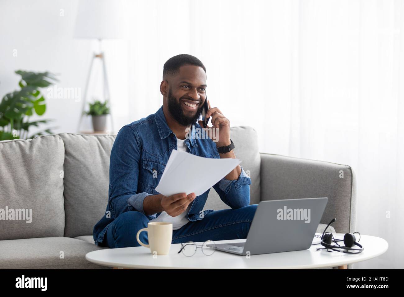 Happy attractive millennial african american bearded guy works with documents and computer, calls by phone Stock Photo