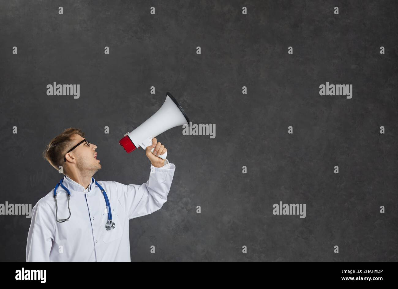 Male healthcare worker loudly making announcement using megaphone while standing on gray background. Stock Photo