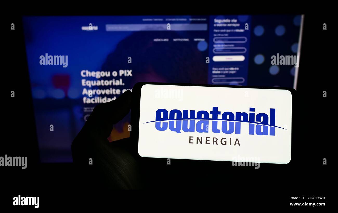 Person holding smartphone with logo of Brazilian energy company Equatorial Energia S.A. on screen in front of website. Focus on phone display. Stock Photo