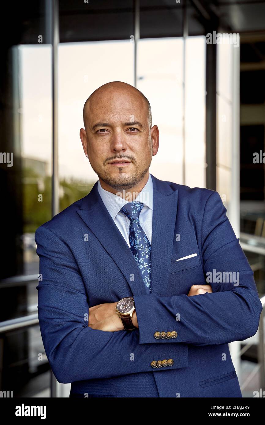 Self assured bald businessman crossing arms and looking at camera while standing against glass wall of modern building Stock Photo