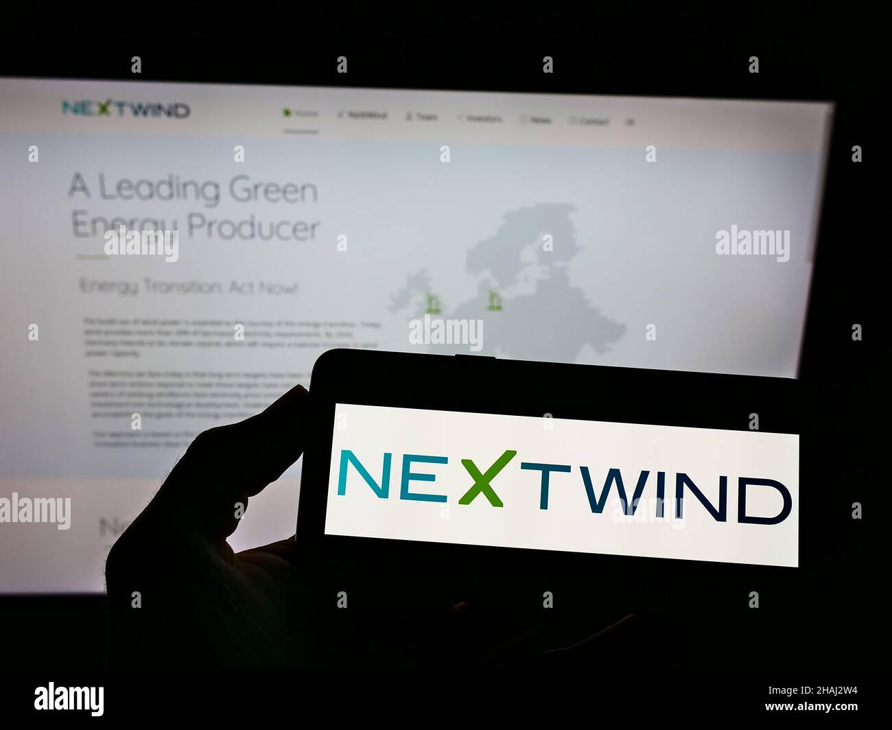 Person holding smartphone with logo of wind energy company NeXtWind Management GmbH on screen in front of website. Focus on phone display. Stock Photo