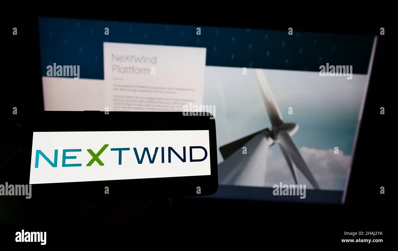 Person holding mobile phone with logo of wind energy company NeXtWind Management GmbH on screen in front of web page. Focus on phone display. Stock Photo