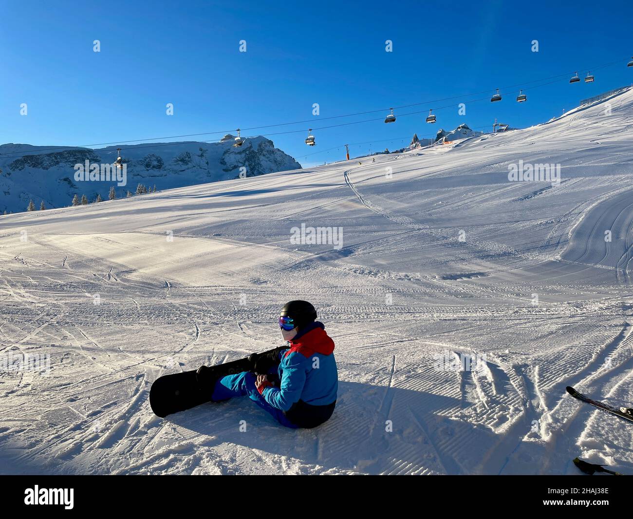 Snowboarder with facial mask during Corona pandemic sitting on snow on a beautiful winter day in winter resort Golm, Montafon, Austria. Stock Photo