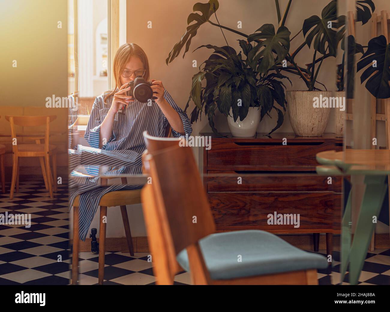 Young woman taking photo of herself, her reflection in mirror in modern cafe with day light and plants. Photographer with professional camera. Stock Photo