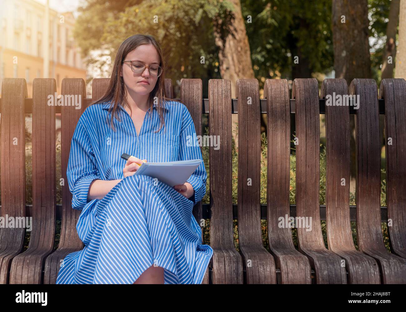 Woman student in glasses sitting on bench in city park with syllabus or notebook with highlighter. Female studying outdoors. Stock Photo