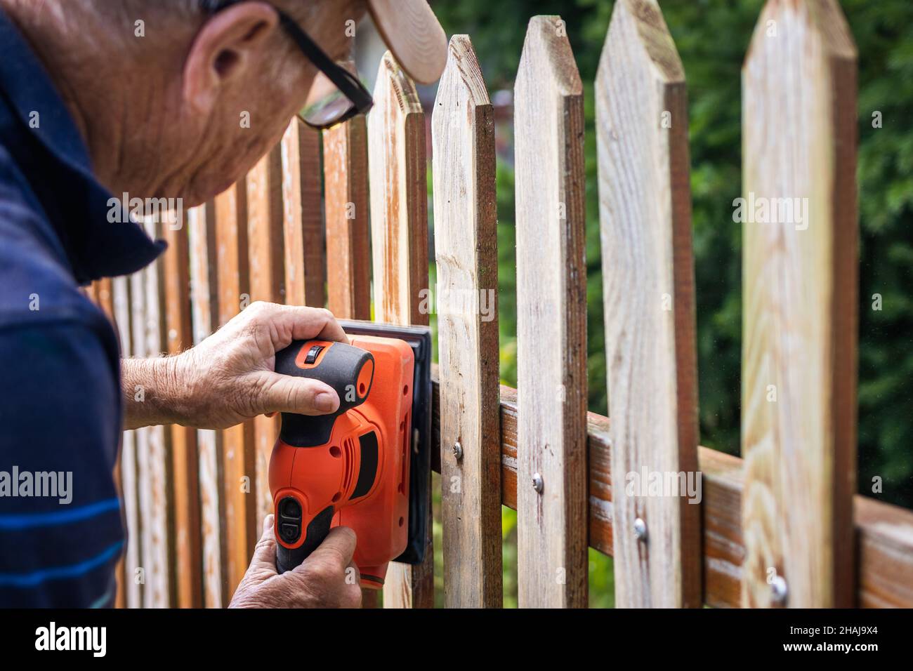 Sanding wooden plank. Senior man grinding picket fence with sander Stock Photo
