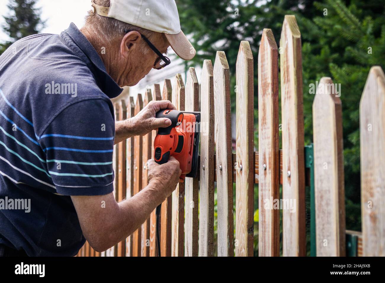 Sanding and preparing wooden fence for painting. Senior man restoring picket fence in garden Stock Photo