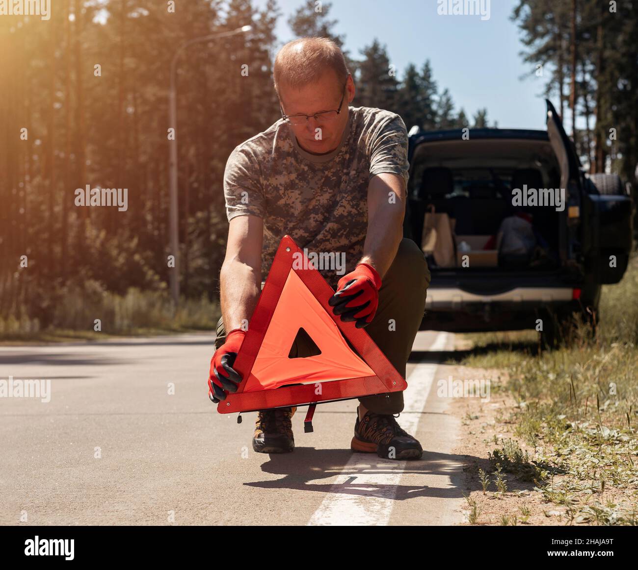 Driver man putting triangle caution sign on road near broken auto in summer. Stock Photo