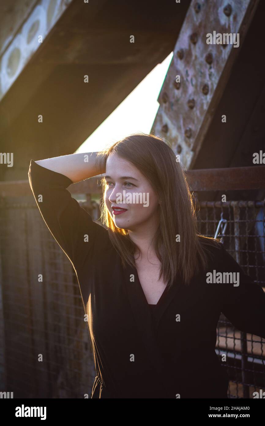 Young beautiful woman standing at bridge and enjoying sunset in city. Fashionable attractive brunette woman outdoors Stock Photo