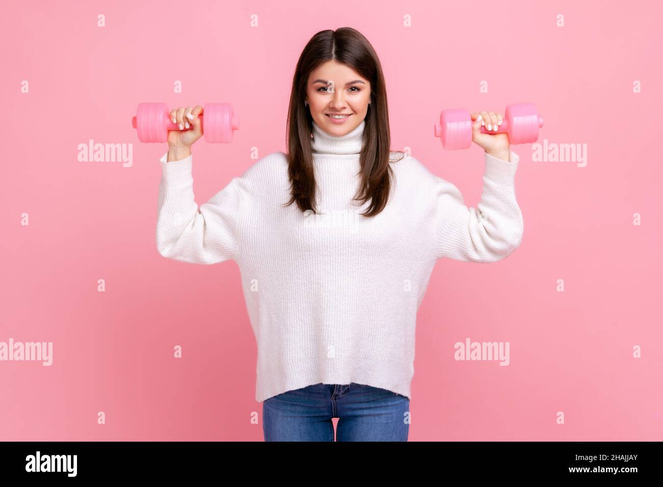 Happy smiling sportive energetic female holding rose dumbbells in raised arms, healthy lifestyle, wearing white casual style sweater. Indoor studio shot isolated on pink background. Stock Photo