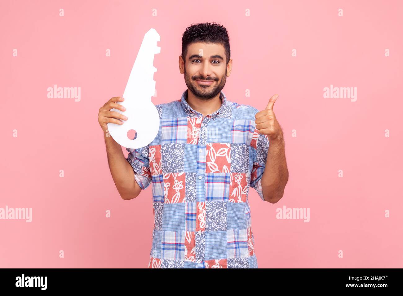 Portrait of positive handsome man in blue shirt holding huge paper key and smiling satisfied with new home, showing thumb up, rental service. Indoor studio shot isolated on pink background. Stock Photo