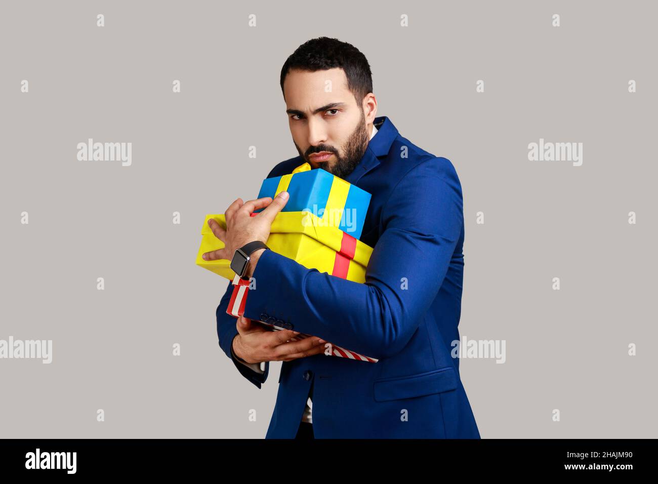 Portrait of angry serious bearded man holding stack of presents in hands, being greedy to share gifts, wearing official style suit. Indoor studio shot isolated on gray background. Stock Photo